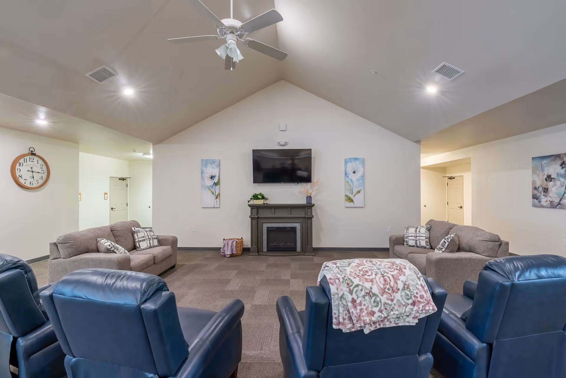 A spacious living room with a vaulted ceiling and ceiling fan. The room features two gray sofas facing each other, and four blue recliner chairs in the foreground, one with a patterned blanket draped over it. On the far wall, there is a flat-screen TV mounted above a dark fireplace mantel, flanked by two floral paintings. The walls are light-colored, and there is a large round clock on the left wall. The room is well-lit with recessed lighting.