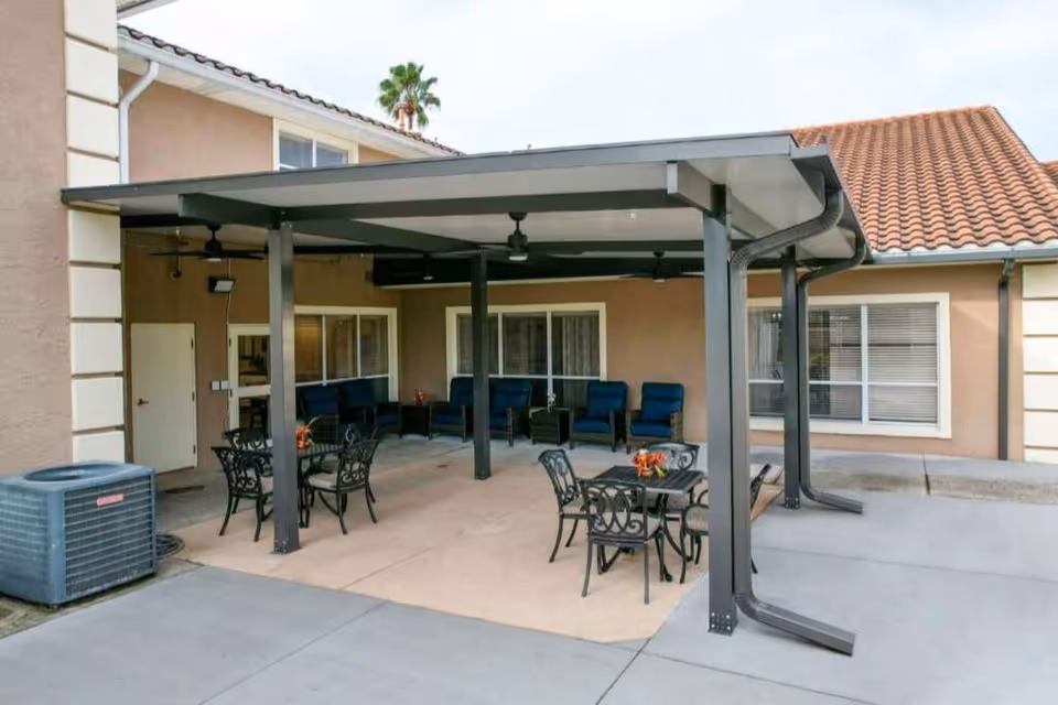 Outdoor covered patio area with metal tables and chairs, cushioned seating along the wall, ceiling fans, and a tiled roof building in the background.