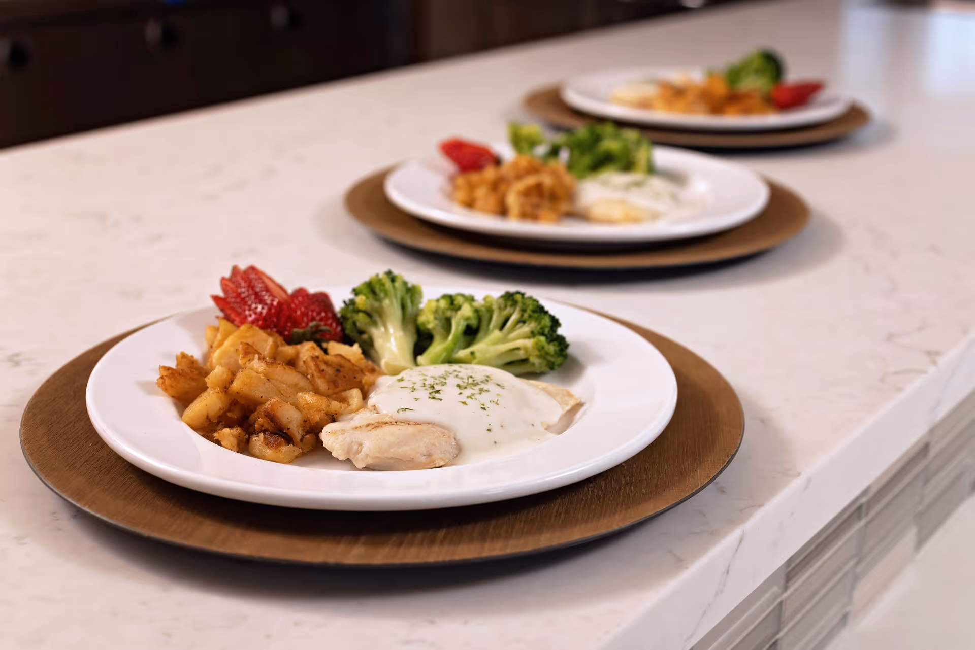 Three white plates with a meal consisting of cooked chicken breast with white sauce, steamed broccoli, diced cooked potatoes, and sliced strawberries, arranged in a row on a white marble countertop.