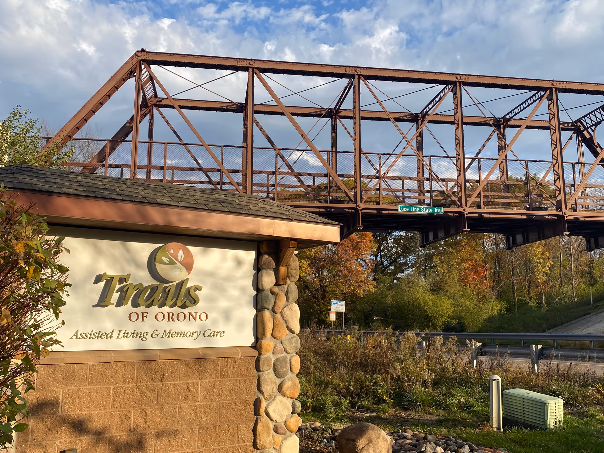 Outdoor scene showing a sign for Trails of Orono Assisted Living & Memory Care in front of a stone and brick structure, with a rusty metal bridge and trees with autumn foliage in the background under a partly cloudy sky.