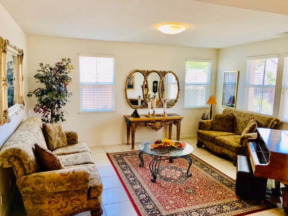 Bright traditional living room with patterned sofas, a glass coffee table on a red area rug, an ornate console table with mirrors, and a piano.