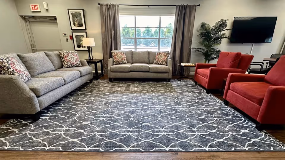 Spacious lounge with gray sofas, two red armchairs, a patterned rug, side tables and a wall-mounted TV beneath a large window.