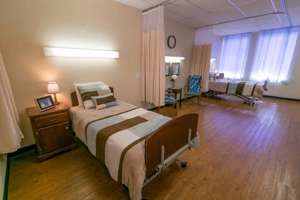 A shared hospital-style room with two beds separated by curtains. Each bed has a wooden headboard and footboard, with beige and brown bedding. There are bedside tables with lamps and framed pictures, chairs, and a clock on the wall. The room has wooden floors and large windows with sheer curtains letting in natural light.