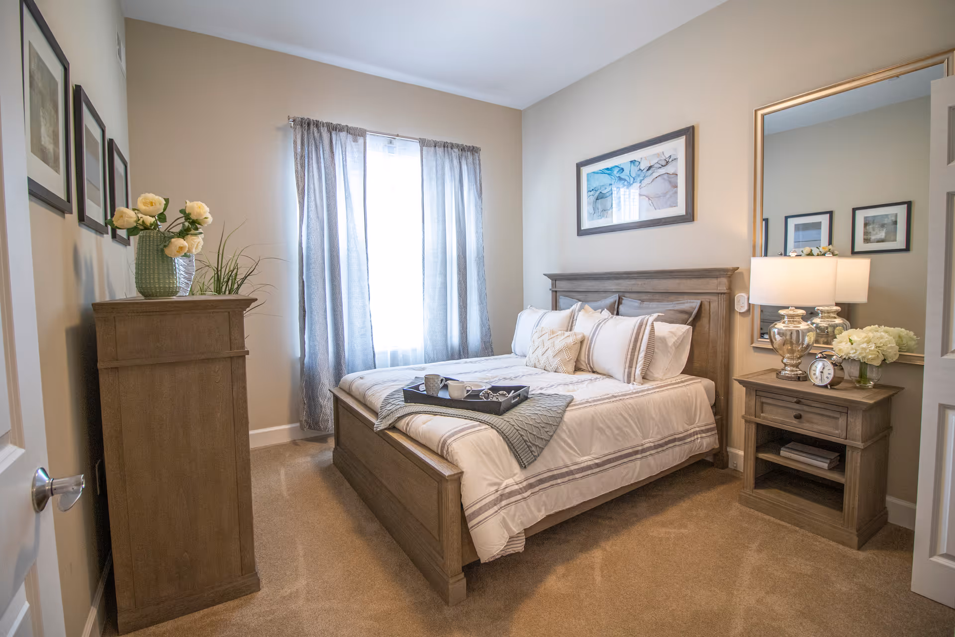 A neatly staged bedroom with a wooden bed, matching nightstand and dresser, neutral decor and a curtained window.