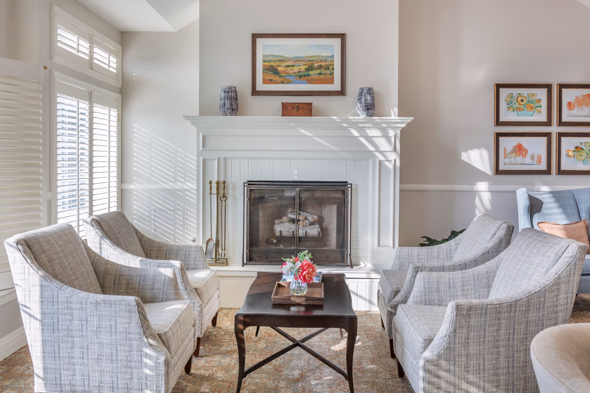 A cozy living room area with four light gray upholstered armchairs arranged around a dark wooden coffee table with a small flower arrangement. The room features a white fireplace with a mesh screen, flanked by fireplace tools. Above the fireplace is a framed landscape painting, and to the right are four framed floral and abstract artworks on the wall. Sunlight streams through white plantation shutters on the left, casting shadows on the walls.