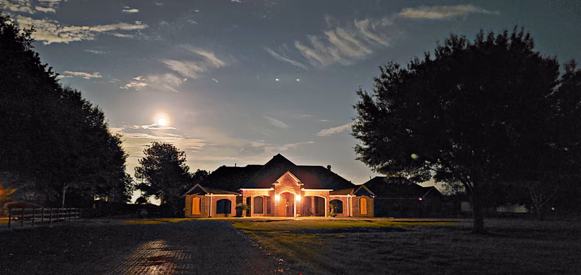 A large house illuminated by exterior lights at night with a full moon and scattered clouds in the sky. The house is surrounded by trees and a grassy area.