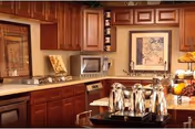 A warm kitchen interior featuring wooden cabinets, a microwave, a framed picture on the wall, and a countertop with several silver coffee carafes and a fruit bowl.