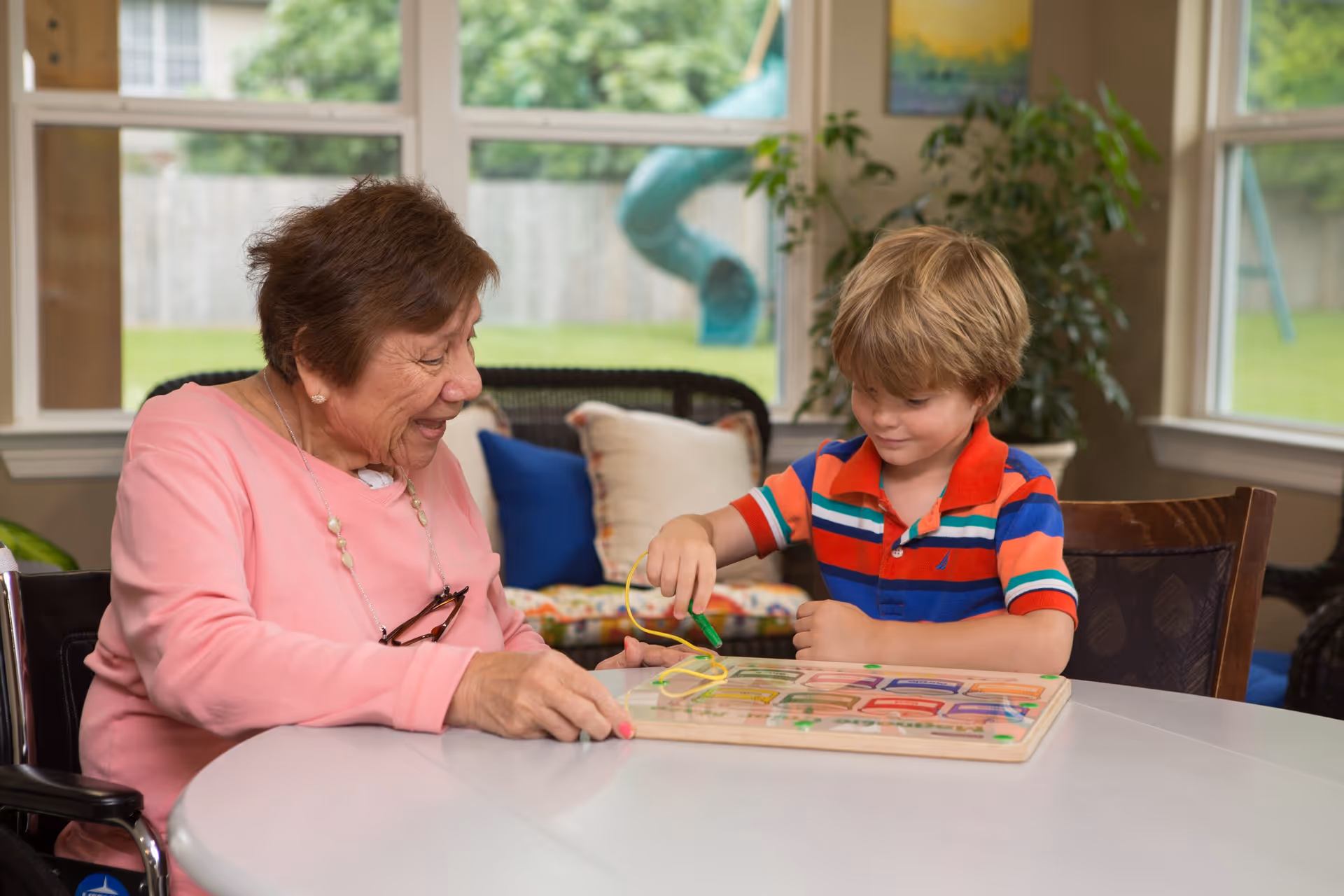An elderly woman and a young boy sitting at a round table playing with a wooden educational toy inside a bright room with large windows showing a green yard outside.