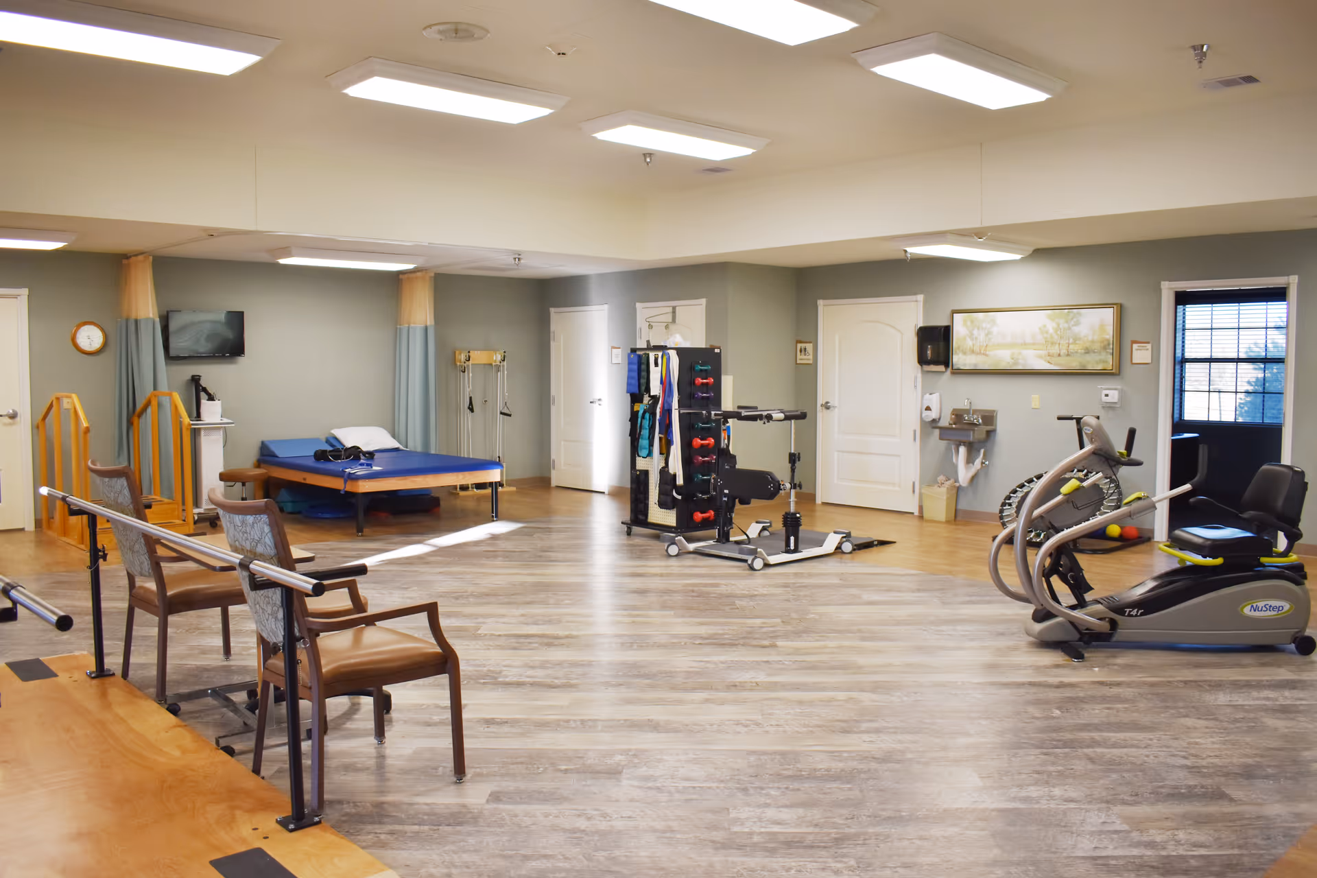 Spacious rehabilitation room with parallel bars, exercise machines, a treatment table, and chairs.