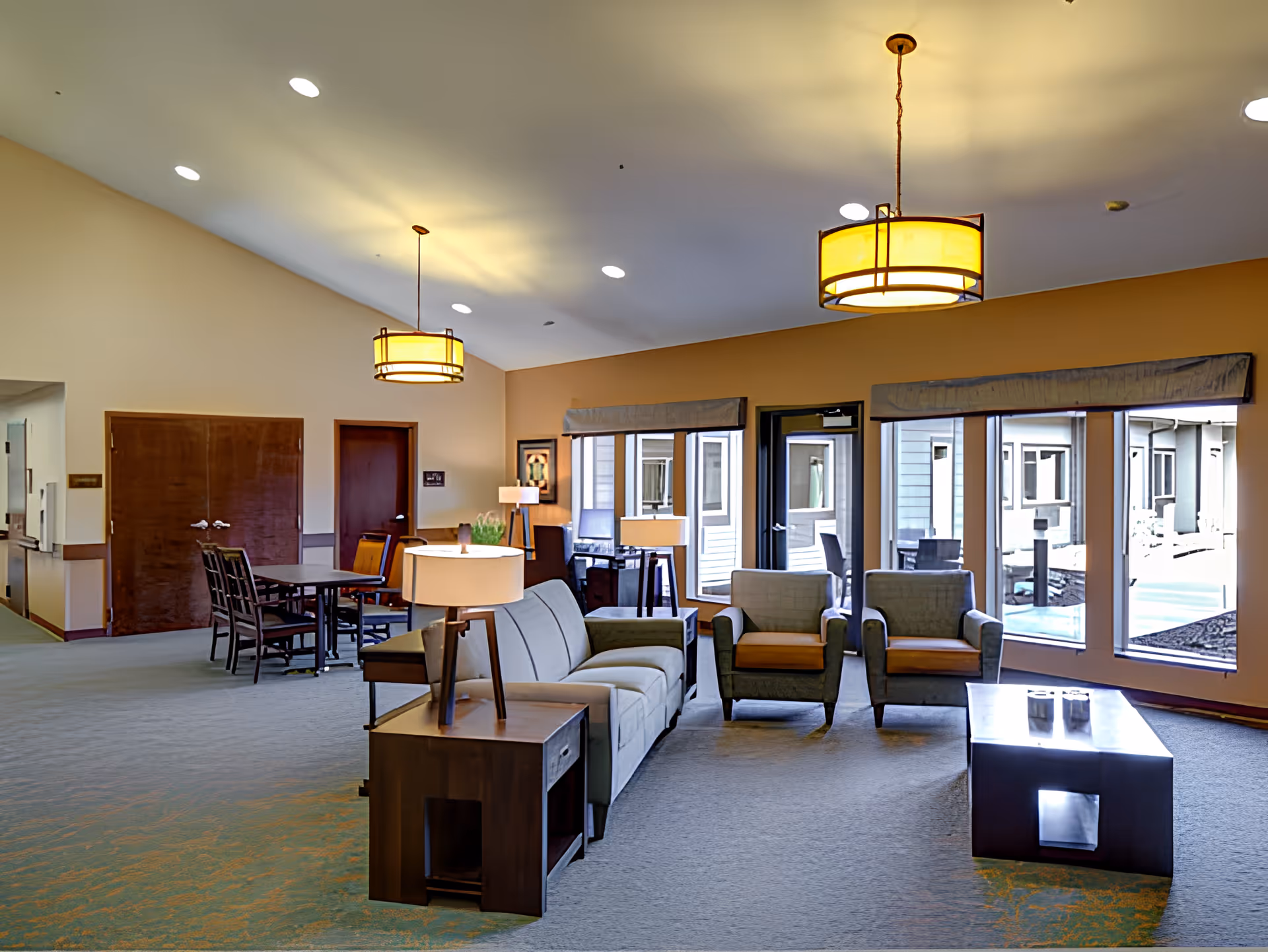 A spacious and well-lit common area with a sofa, two armchairs, a coffee table, and side tables with lamps. Large windows provide a view of an outdoor patio area. There is also a dining table with chairs and warm ceiling lights hanging from the high ceiling.