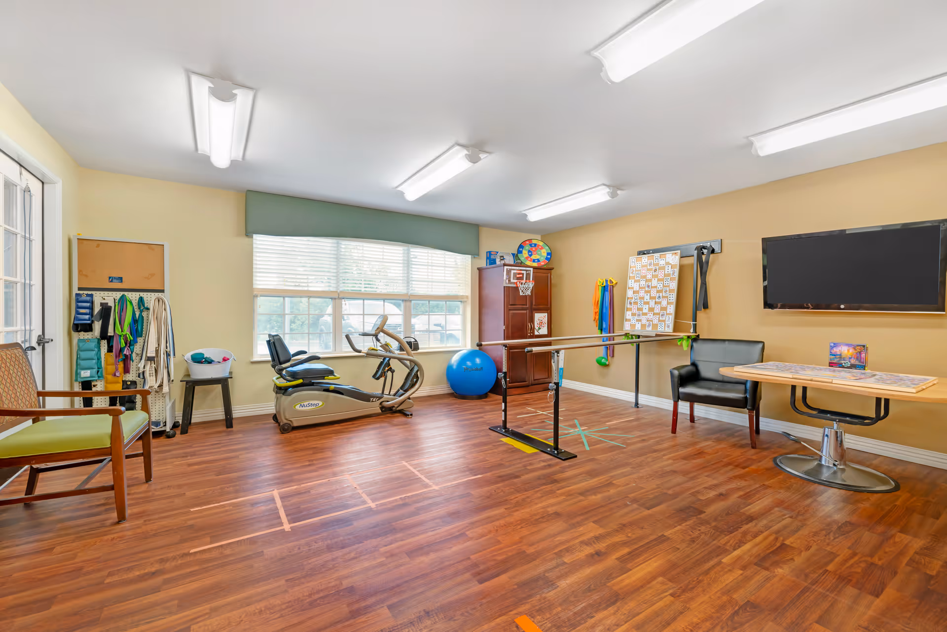 Bright multipurpose exercise/activity room with an exercise bike, therapy parallel bars, exercise ball, table and chairs.