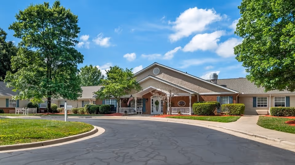 Exterior view of Brookdale South Park facility showing a single-story building with a covered entrance, surrounded by green trees and bushes under a blue sky with some clouds. There is a curved driveway in front of the entrance.