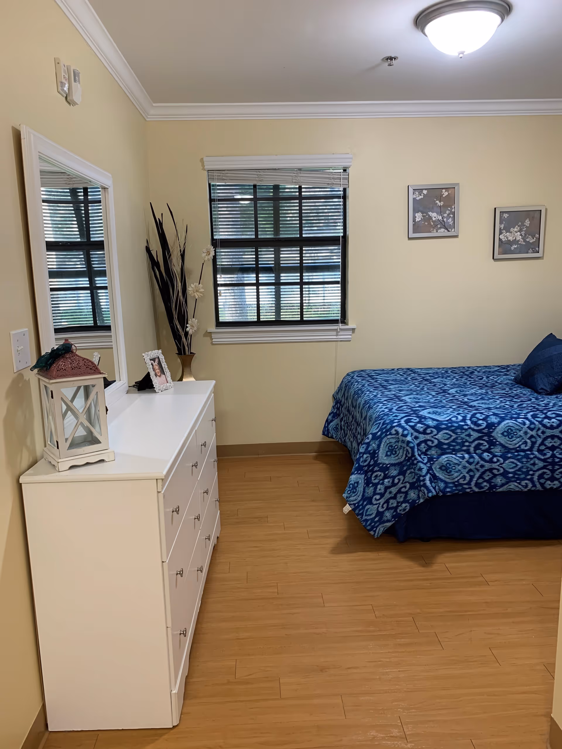 A bedroom with light yellow walls and wooden flooring. There is a white dresser with a mirror on the left side, decorated with a lantern, a framed photo, and a vase with tall decorative sticks and flowers. A window with closed blinds is centered on the back wall. On the right side, there is a bed with a blue patterned bedspread and matching pillows. Two framed floral artworks hang on the wall above the bed. The ceiling has a round light fixture.
