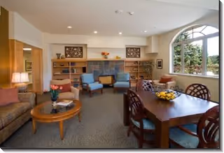 A cozy living room area in Bay Vista Commons featuring a fireplace with a wooden mantel, two blue armchairs, a brown sofa, a round wooden coffee table with flowers, and a wooden dining table with chairs. Large windows allow natural light to brighten the room.