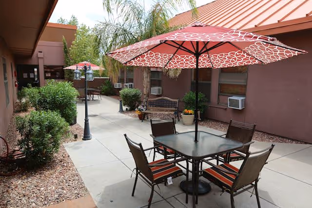 Outdoor patio area at Coronado Healthcare Center with tables and chairs under red patterned umbrellas, surrounded by plants and bushes along a paved walkway next to the building.
