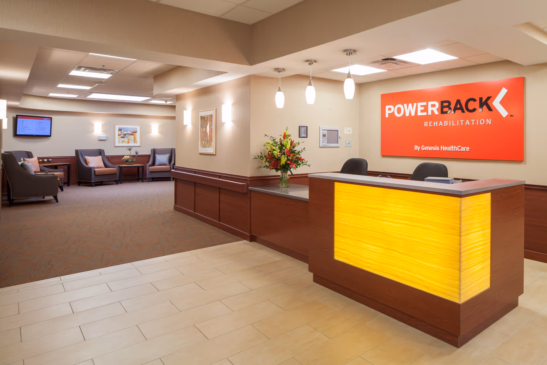 Reception area of a rehabilitation facility with a wooden desk featuring a yellow illuminated front panel. Behind the desk is a red sign that reads 'POWERBACK REHABILITATION By Genesis HealthCare'. To the left, there is a seating area with several armchairs, a small table with a flower arrangement, and framed artwork on the walls. The floor is tiled near the desk and carpeted in the seating area. The ceiling has recessed lighting and pendant lights above the desk.