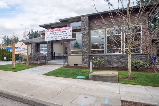 Exterior view of Senior Haven RCF building with a sidewalk and small landscaped area in front. A sign near the sidewalk displays the facility name and contact information. A banner above the entrance announces a grand opening in 2018 and mentions new rooms and revenue acceptance.