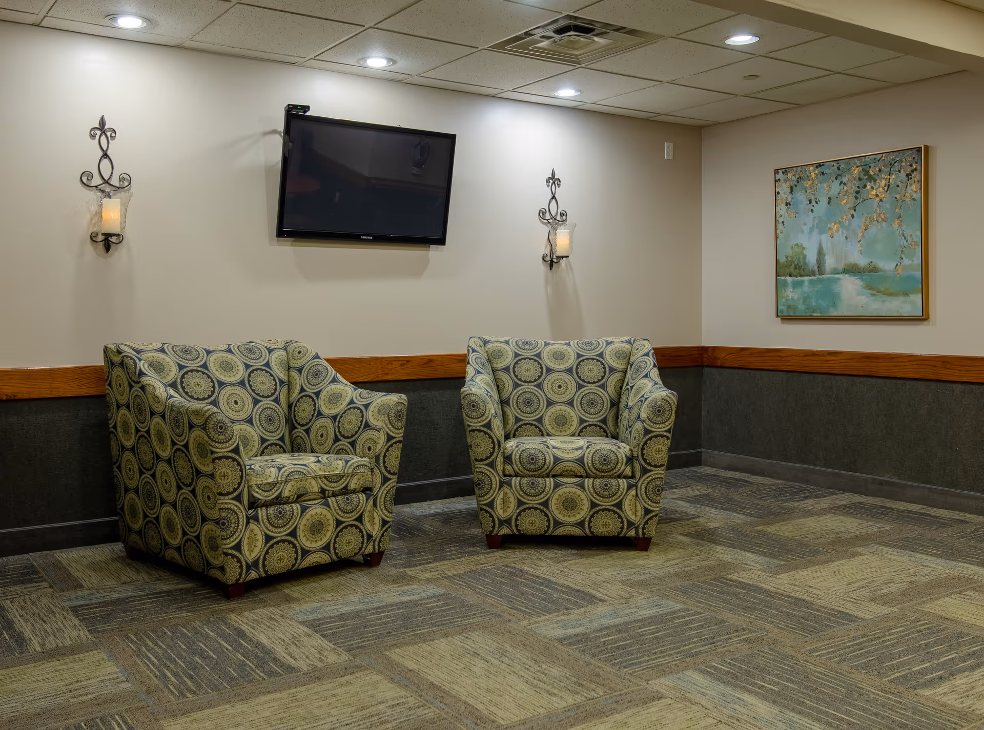 A small sitting area with two patterned armchairs placed side by side against a beige wall. Above the chairs is a wall-mounted flat screen TV. On either side of the TV are decorative wall sconces with candles. To the right, there is a framed painting depicting a serene landscape with trees and water. The floor is carpeted with a patterned design in shades of green and brown.