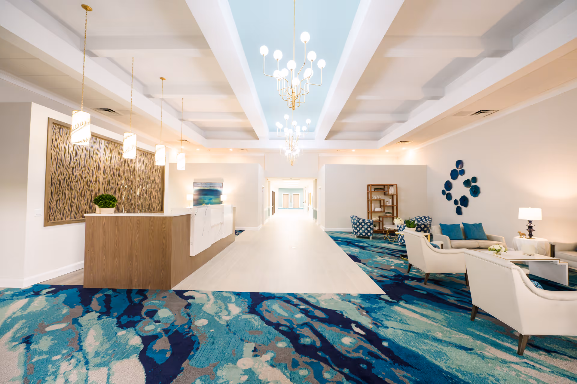 Bright and spacious interior lobby area with a wooden reception desk on the left, modern pendant lights hanging from the ceiling, and a seating area with white chairs and blue cushions on the right. The floor has a vibrant blue and teal patterned carpet, and the ceiling features white beams with a light blue inset and chandeliers.