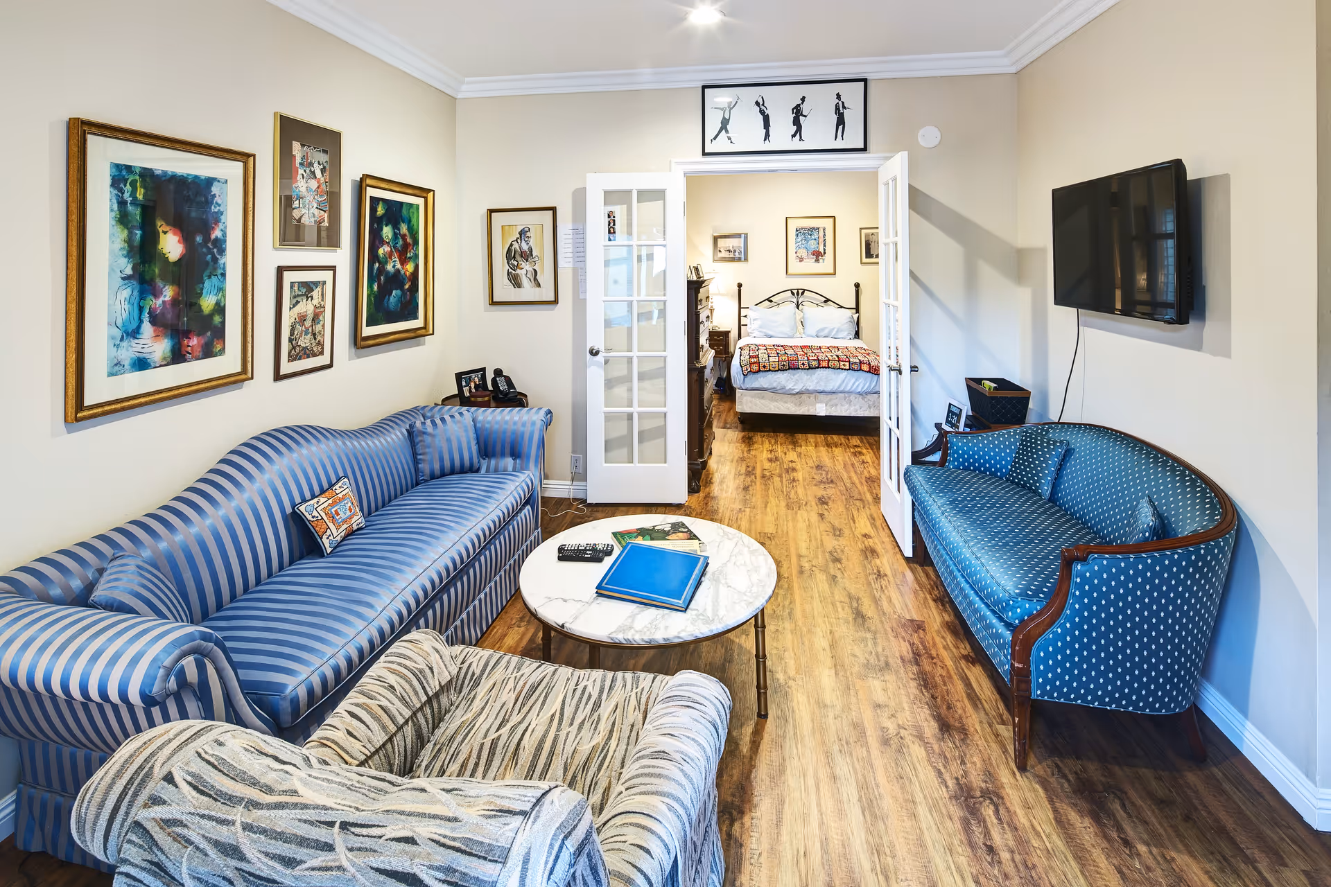 A cozy living room with a blue striped sofa, a patterned armchair, and a blue polka dot loveseat. A round marble coffee table with books and a remote control sits in the center. The walls are decorated with framed artwork. French doors open to a bedroom with a bed, nightstand, and dresser. A flat-screen TV is mounted on the wall to the right.