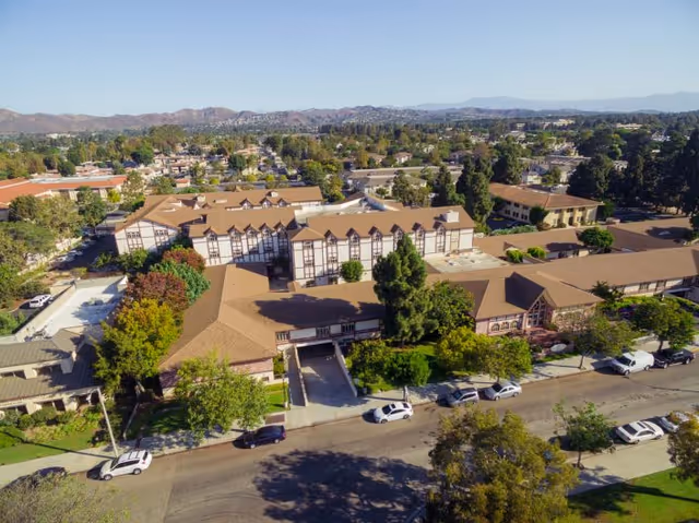 Aerial view of a multi-building assisted living complex with brown roofs, trees, and parked cars along the street.