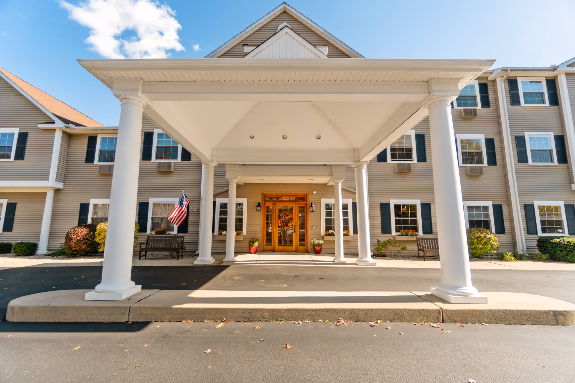 Front entrance of a senior living facility with a covered driveway supported by white columns, beige siding, multiple windows with dark shutters, an American flag, benches, and potted plants near the wooden double doors.