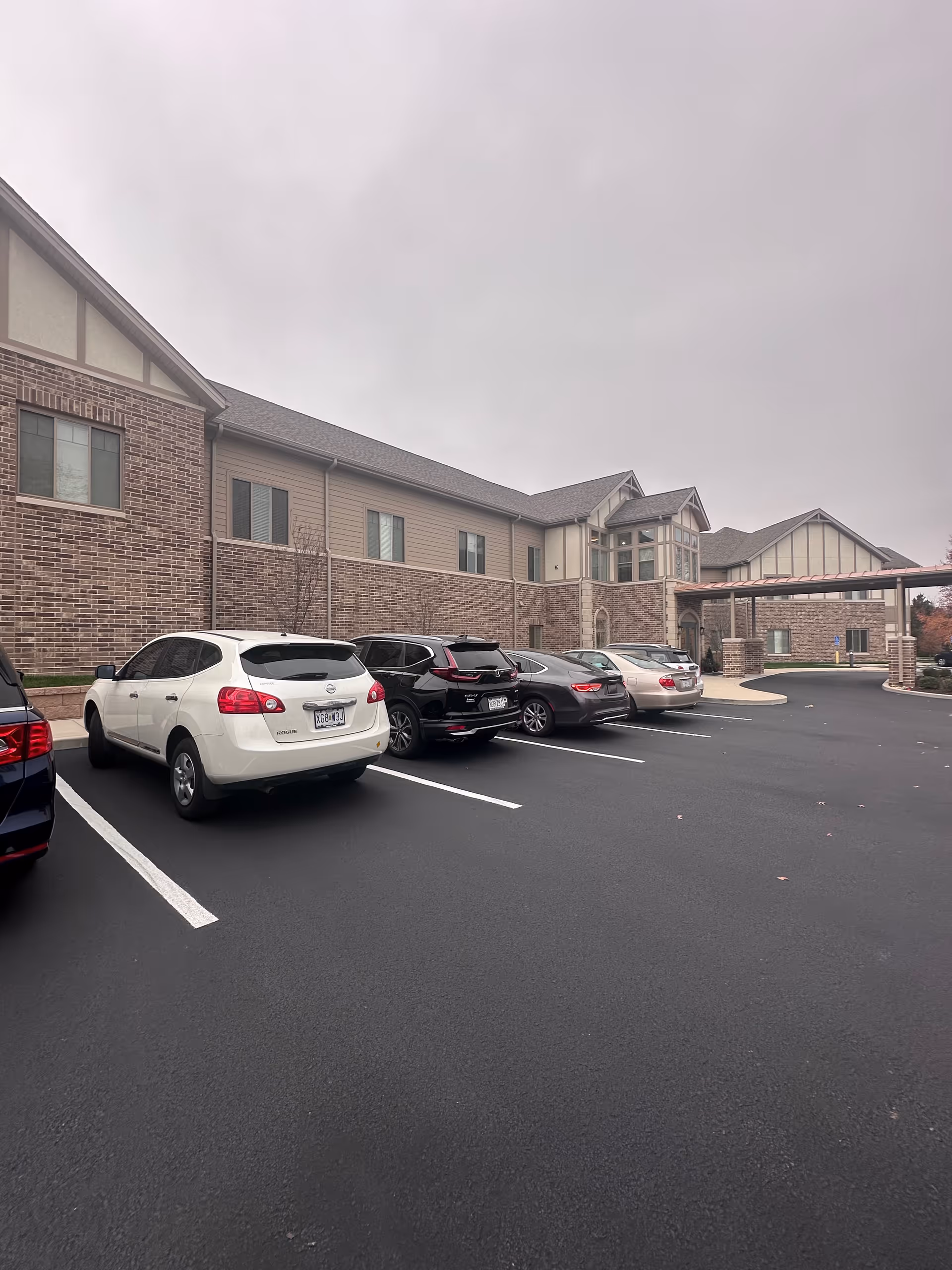 Parking lot with several cars parked in front of a two-story brick senior living building under a gray sky.