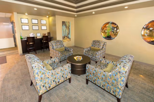 A cozy seating area in a senior living facility with four patterned armchairs arranged around a round coffee table. The room has beige walls with two circular windows showing an aquarium behind them. There is a small counter with two high chairs and framed pictures on the wall in the background.