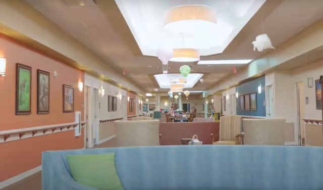 A long, well-lit hallway in a senior living facility with comfortable seating areas including a blue couch with a green pillow and beige armchairs. The walls are decorated with framed artwork and the ceiling features hanging light fixtures and decorative hot air balloon ornaments. The hallway has doors on both sides and a warm, inviting atmosphere.