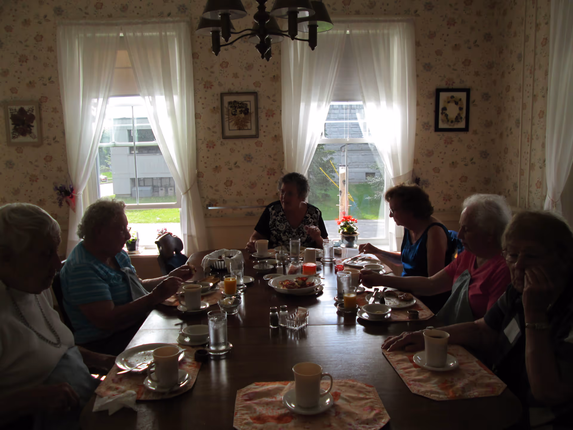 A group of elderly women sitting around a wooden dining table having a meal together in a room with floral wallpaper and two large windows with white curtains.