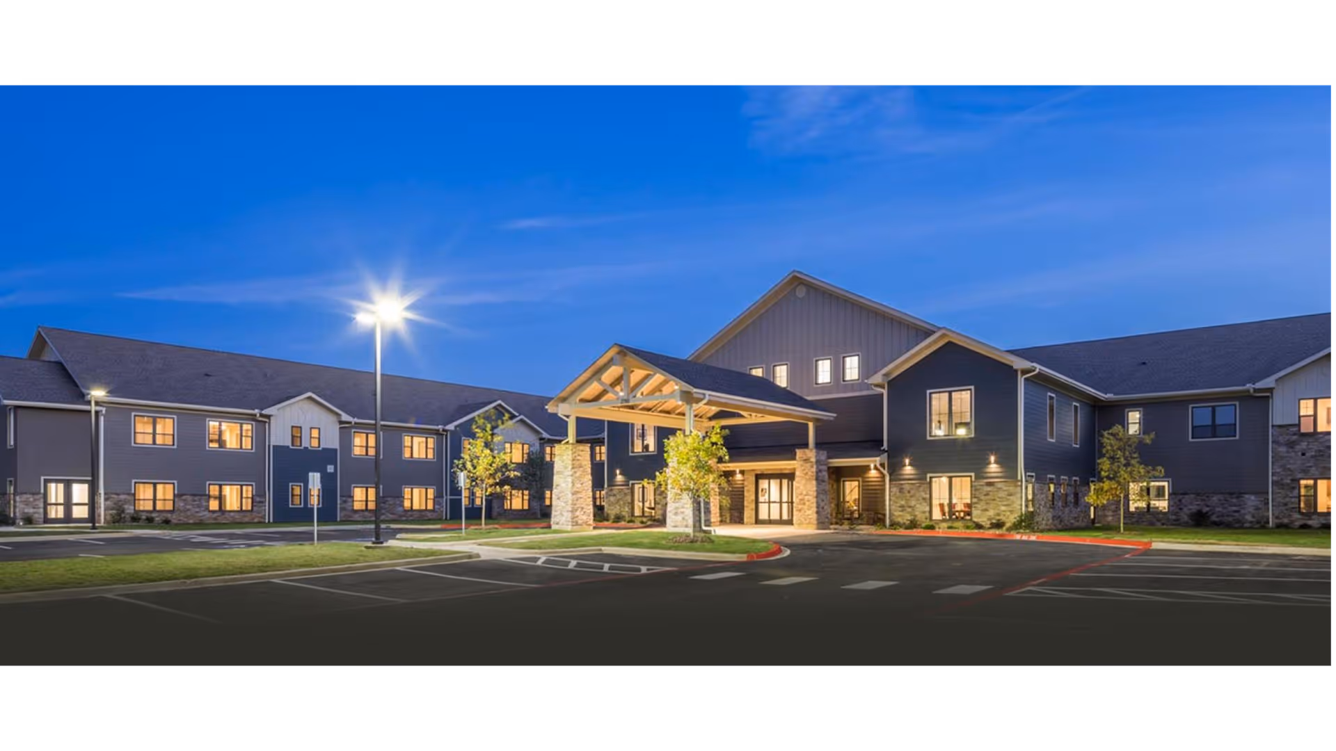 Exterior view of Primrose Retirement Community of Tyler building at dusk with lights on, featuring a covered entrance supported by stone pillars, multiple windows, and a parking area with street lamps and small trees.