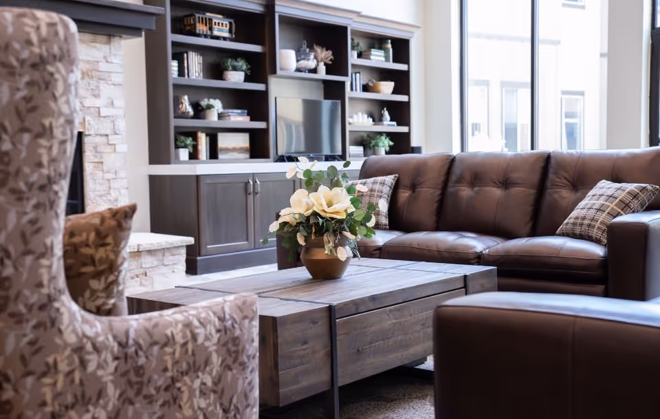 A cozy senior living room featuring a brown leather sofa with plaid pillows, a wooden coffee table with a flower arrangement, a patterned armchair, and a built-in dark wood shelving unit with a TV and decorative items. Large windows allow natural light to fill the space.