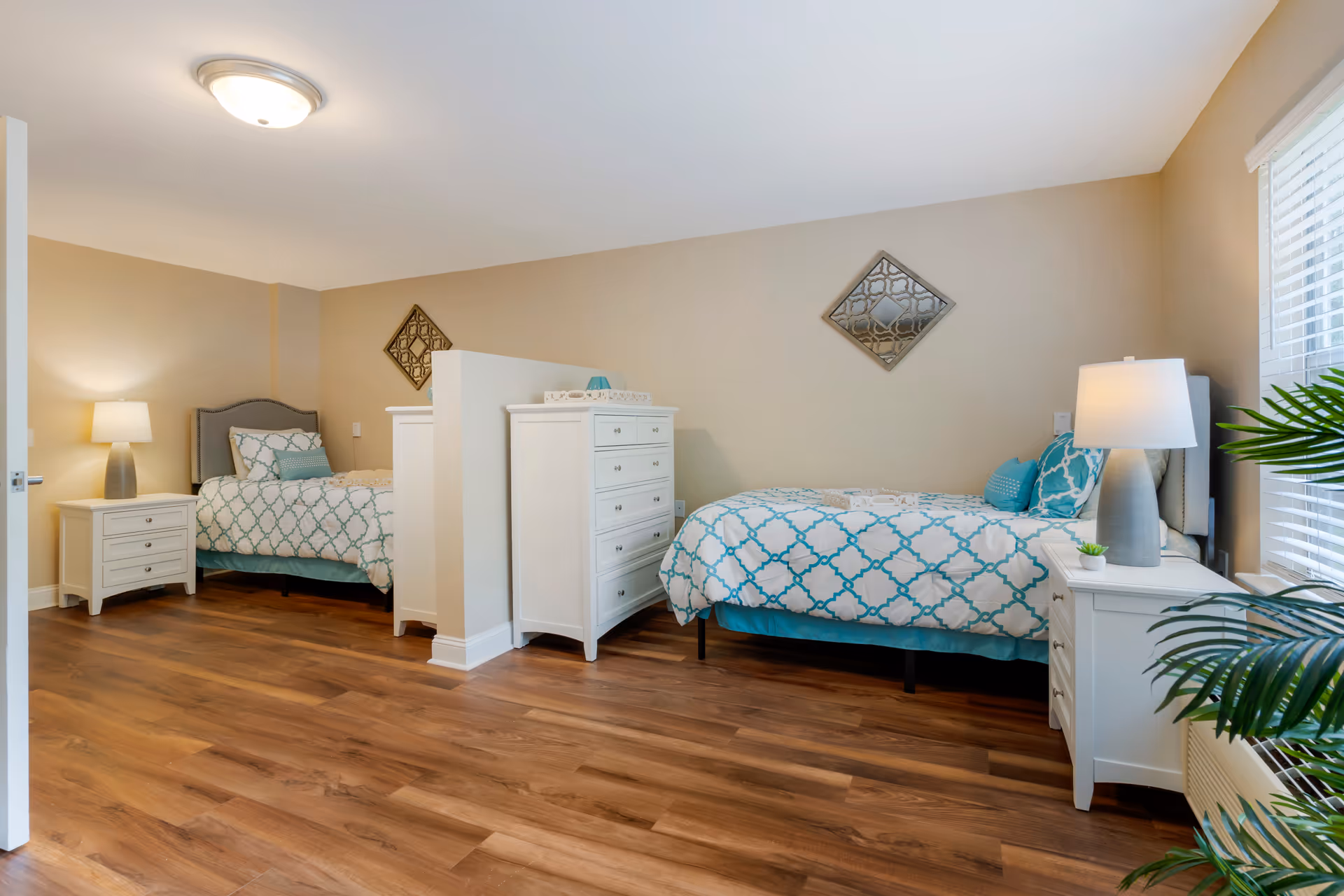 Bright shared bedroom with two twin beds separated by a low partition, white dressers and nightstands, lamps, and wood flooring.