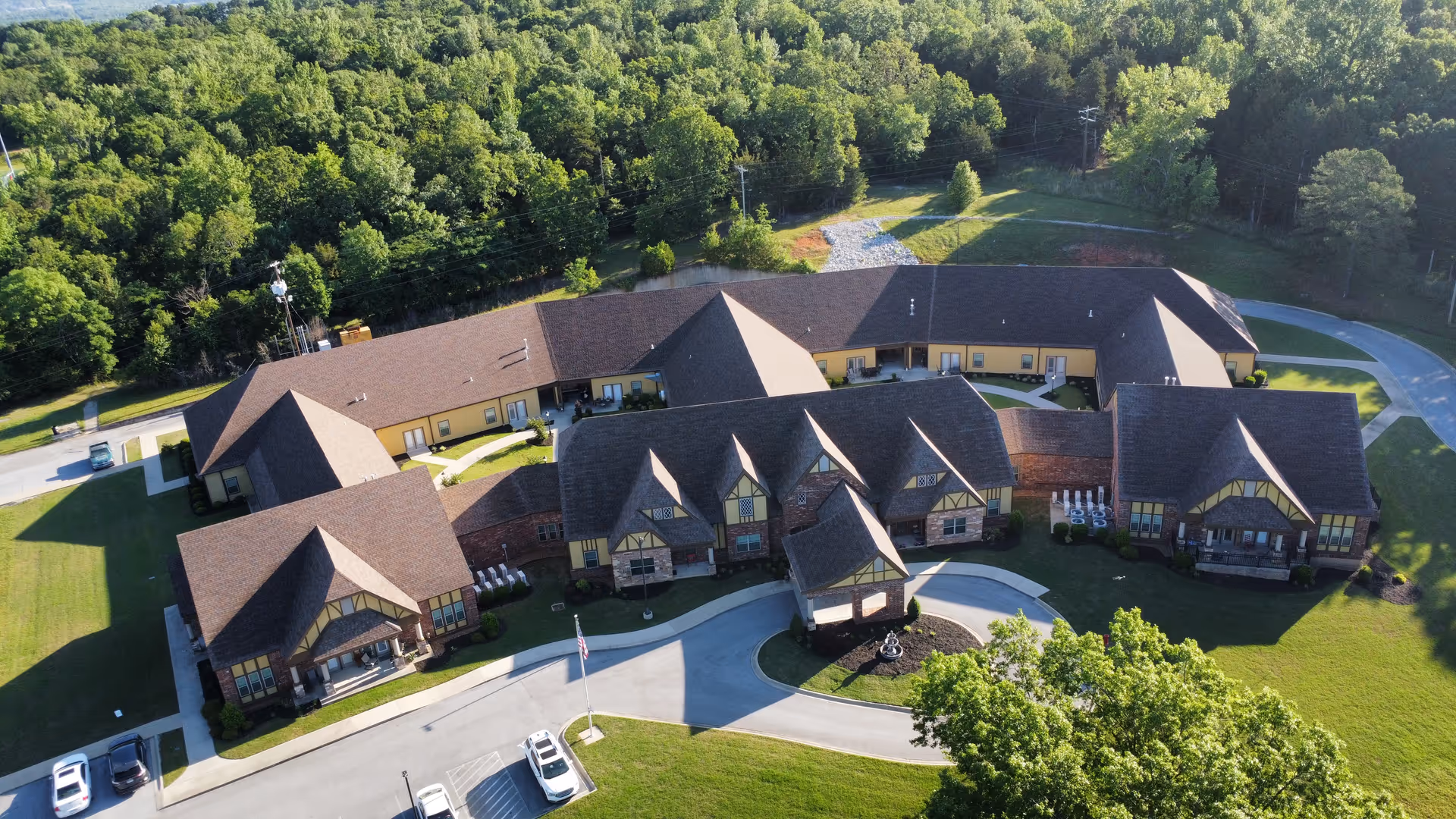Aerial view of Woodcrest Assisted Living facility showing a large, multi-wing building with pitched roofs surrounded by green lawns and trees. There is a circular driveway with parked cars and an American flag near the entrance.