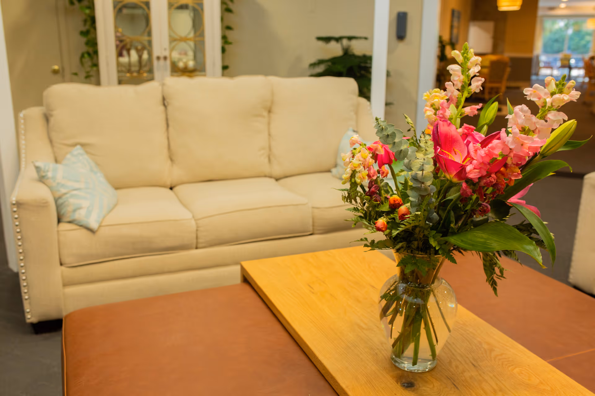 A beige sofa with two light blue patterned pillows in a living room setting. In front of the sofa is a wooden coffee table with a glass vase containing a colorful bouquet of flowers including pink lilies and snapdragons. The background shows part of the room with a cabinet and some greenery.