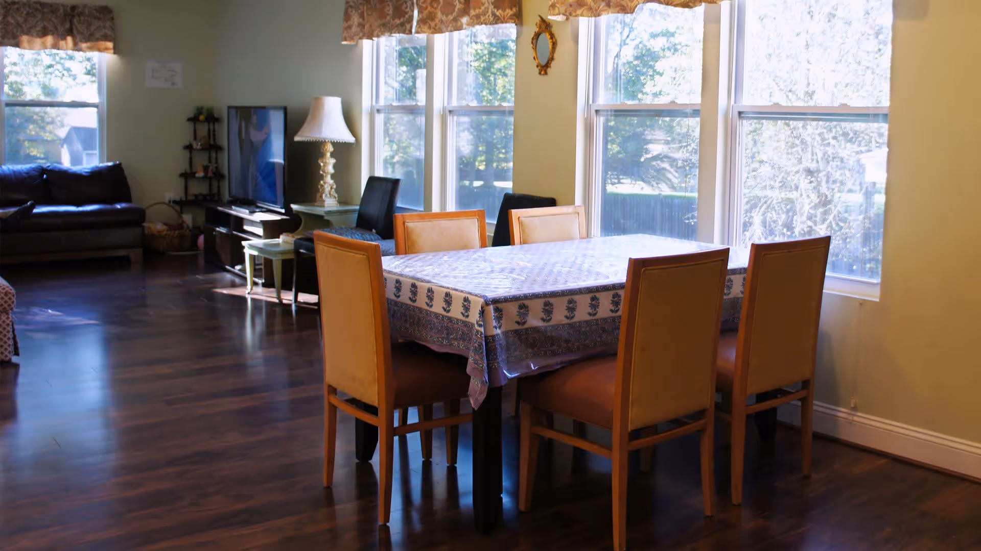 A bright room with large windows letting in natural light, featuring a dining table covered with a patterned tablecloth and surrounded by six chairs. In the background, there is a dark leather sofa, a TV on a stand, a lamp on a side table, and additional seating. The floor is wooden and the walls are light-colored.