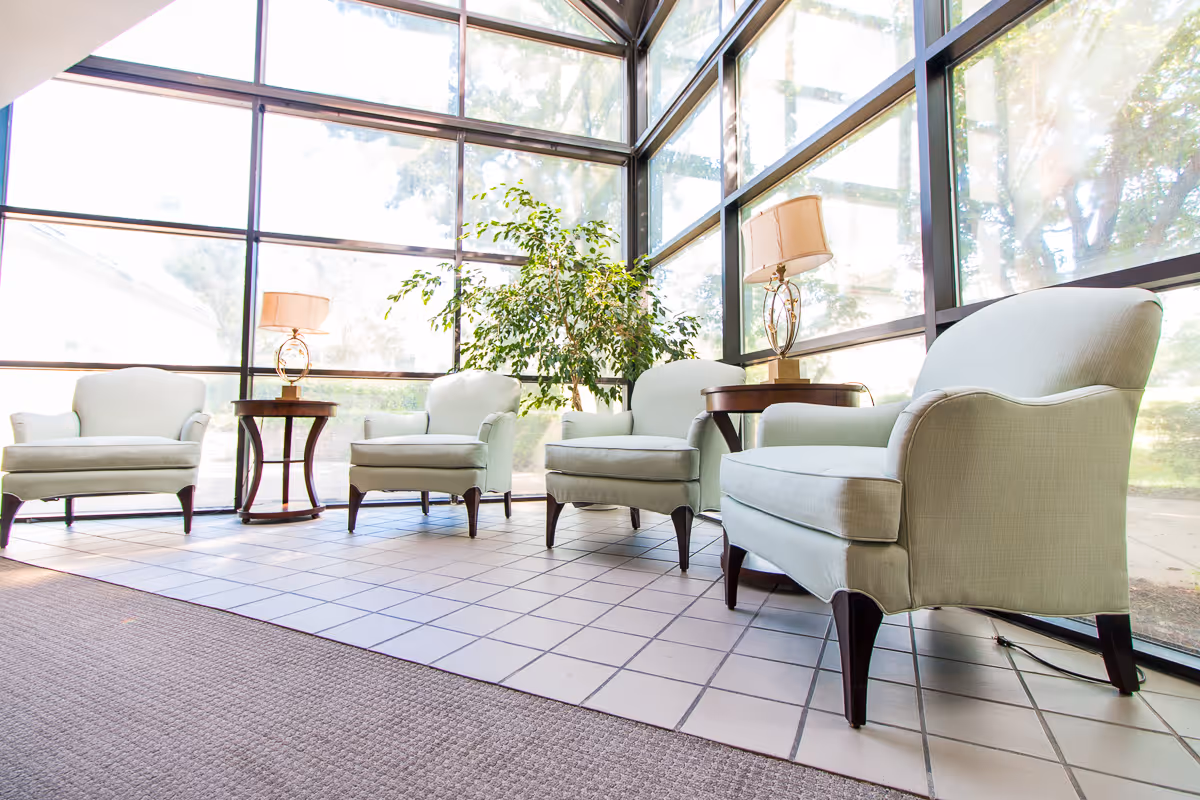A bright seating area with four light-colored upholstered armchairs arranged around two wooden side tables with lamps. The space has large floor-to-ceiling windows letting in natural light and a green plant near the center window.