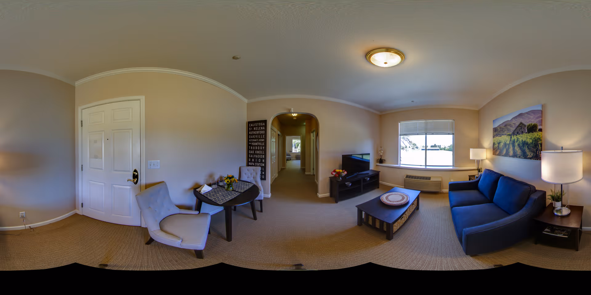 Panoramic view of a furnished living room in a senior living apartment showing a blue sofa, coffee table, TV, small dining table and entry door.