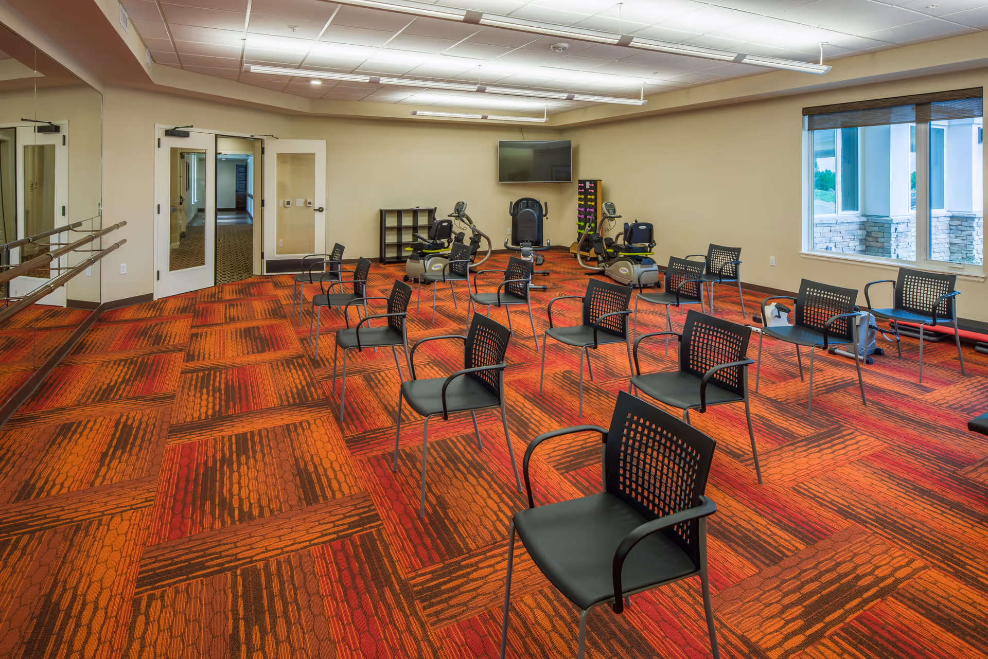 A spacious exercise room with red and orange patterned carpet, several black chairs arranged in rows, exercise equipment including stationary bikes and weights, a large wall-mounted TV, mirrored wall with ballet bars, and a window showing an outdoor view.