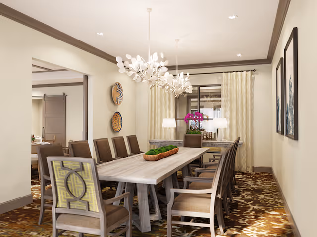 A well-lit dining room with a long wooden table surrounded by ten chairs. The room features two modern white chandeliers hanging from the ceiling, patterned carpet flooring, and light-colored walls with decorative plates and framed artwork. A window with cream-colored curtains and two table lamps is visible at the far end of the room.