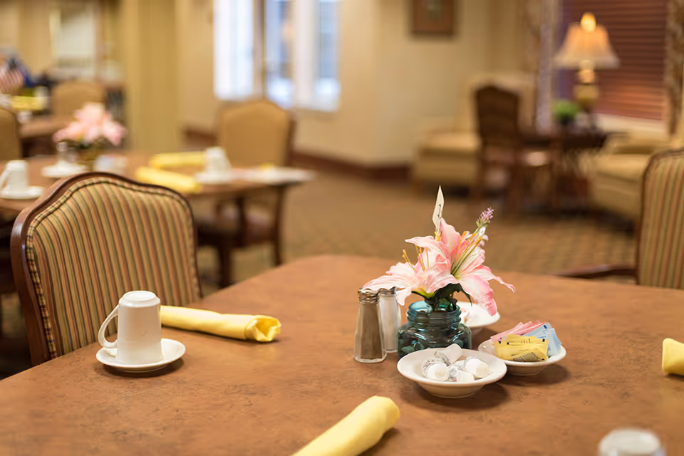 A dining table set with a white coffee cup on a saucer, a yellow folded napkin, salt and pepper shakers, a small vase with pink lilies, and bowls containing creamers and sugar packets. The background shows more tables and chairs in a softly lit dining room.
