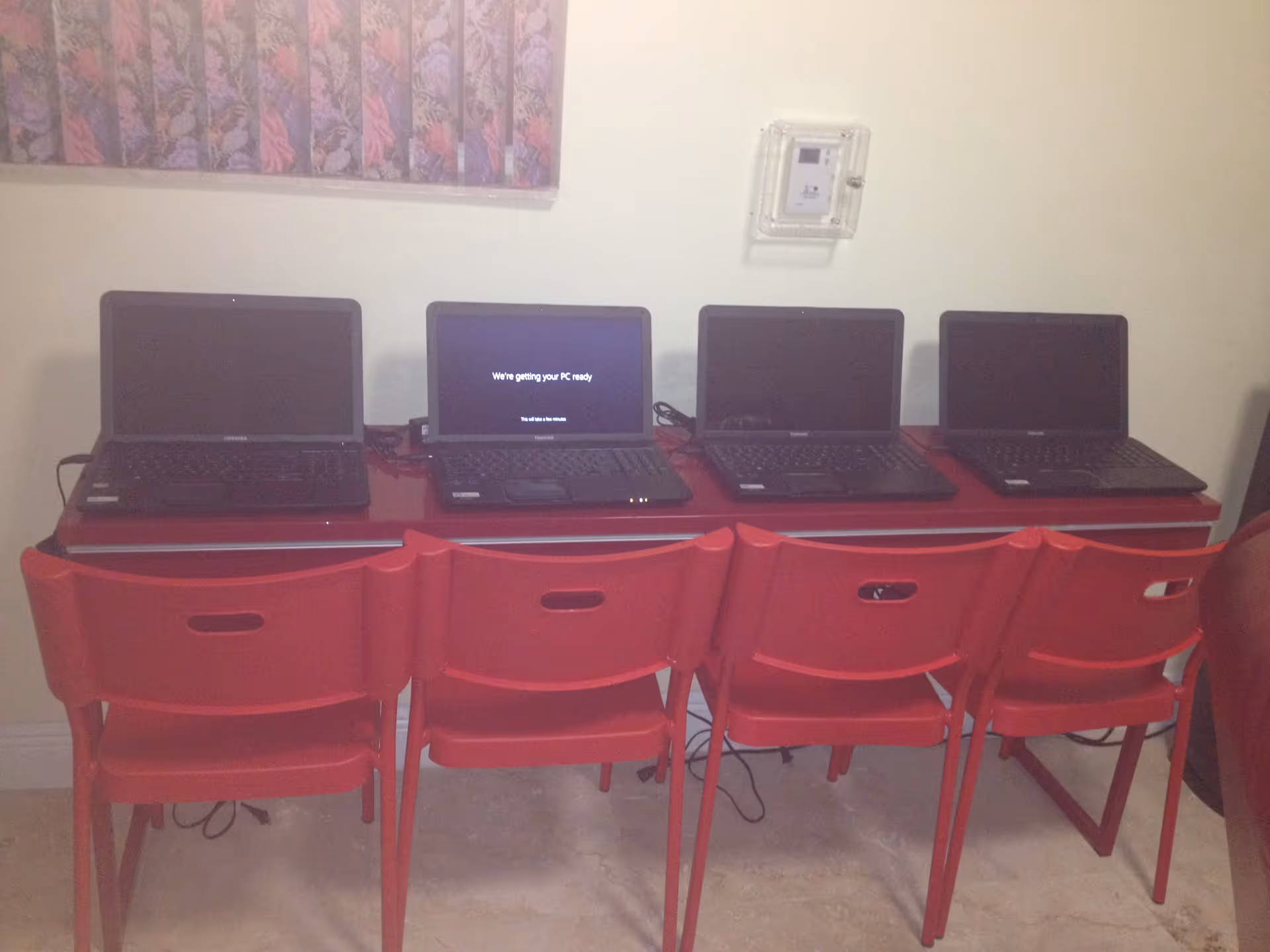 Four black laptops placed on a red table with four red chairs in front of them. One laptop screen displays the message 'We're getting your PC ready'. A floral painting is partially visible on the wall above the laptops.