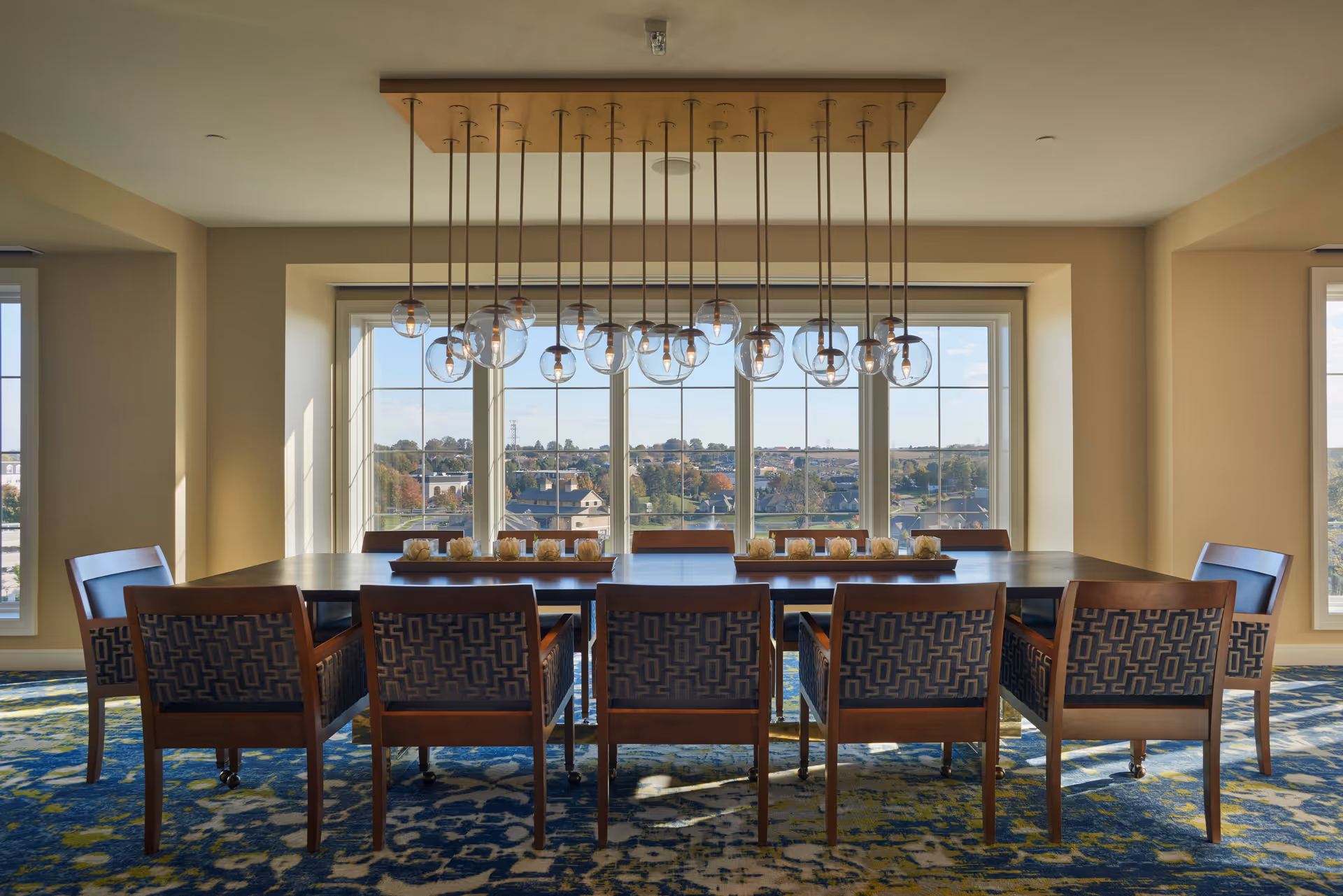 A dining room with a long rectangular wooden table surrounded by eight wooden chairs with patterned upholstery. Above the table hangs a modern chandelier with multiple glass globe light fixtures. Large windows behind the table offer a view of a suburban landscape under a clear sky. The room has beige walls and a blue patterned carpet.