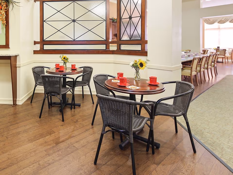 A cozy dining area with two round wooden tables, each surrounded by four black wicker chairs. Each table has a small vase with sunflowers and red coffee cups placed on coasters. The room has wooden flooring and decorative glass panels with wooden frames on the walls. In the background, there is a larger dining area with a long table and multiple chairs near large windows with blinds.