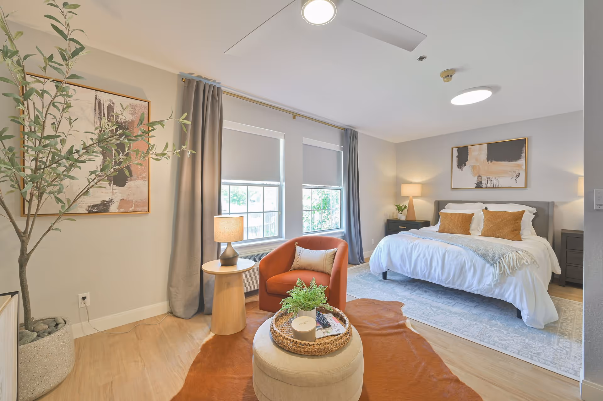 A bright and cozy bedroom with a large bed featuring white bedding and two brown pillows. There are two nightstands with lamps on either side of the bed. A comfortable orange armchair with a small pillow sits next to a round wooden side table with a lamp. In front of the chair is a round ottoman with a woven tray holding a plant, a candle, and a remote control. The room has two large windows with gray curtains and roller shades, light wood flooring, a large potted plant, and abstract artwork on the walls.