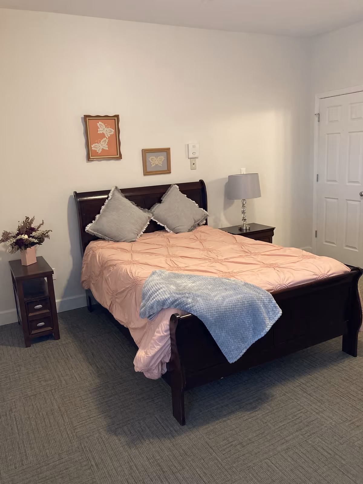 A bedroom with a dark wooden bed frame, a pink comforter, two gray pillows, and a light blue blanket draped over the foot of the bed. There is a nightstand with a lamp on the right side of the bed and a small table with a flower arrangement on the left side. Two framed butterfly artworks hang on the white wall above the bed. A closed white door is visible on the right side of the room.