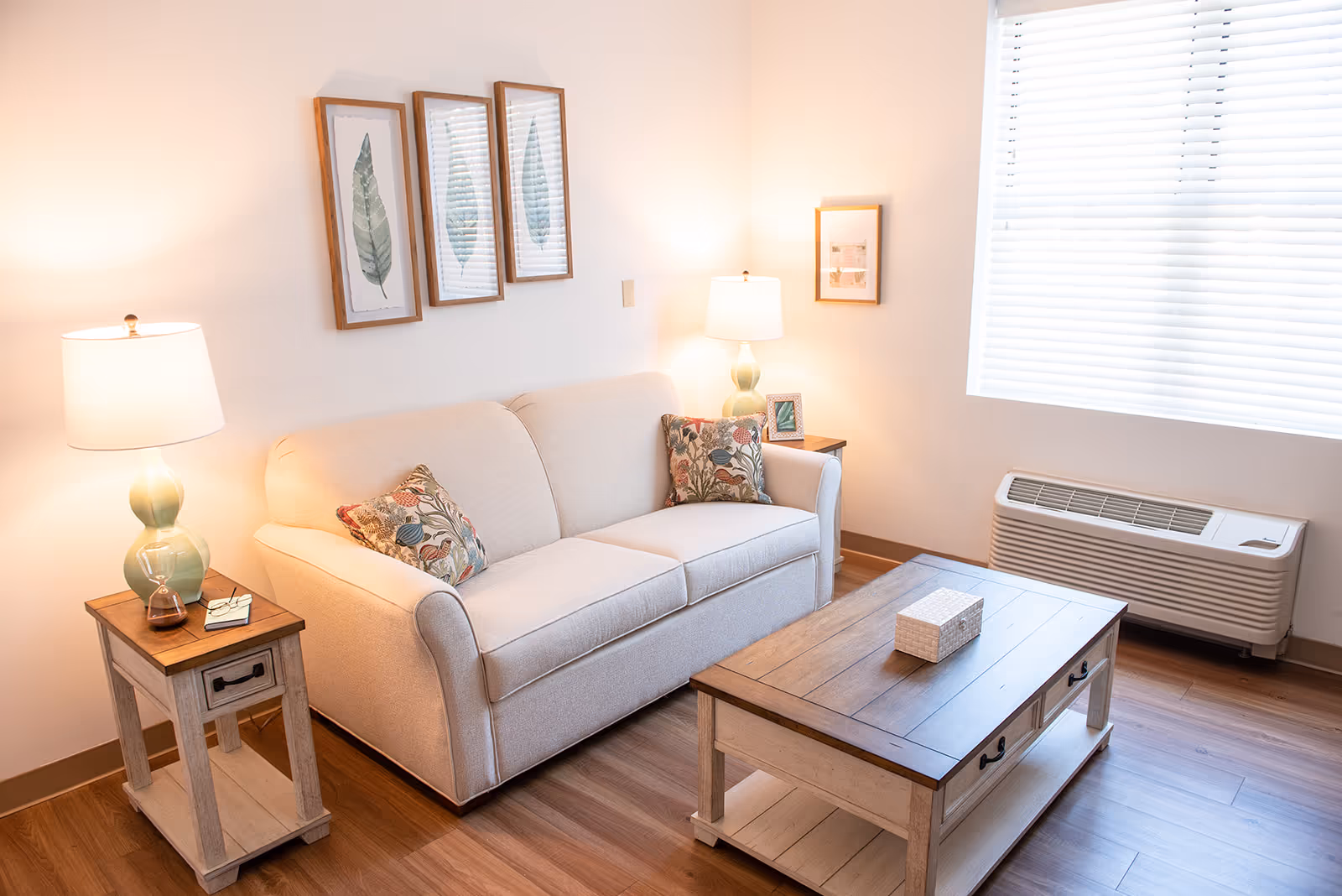 A cozy living room with a beige sofa adorned with two floral patterned cushions. On either side of the sofa are wooden side tables with lamps, one holding a small hourglass and a pair of glasses. Above the sofa are three framed botanical prints. A wooden coffee table with drawers sits in front of the sofa, and a window with white blinds lets in natural light. A wall-mounted air conditioning unit is beneath the window.