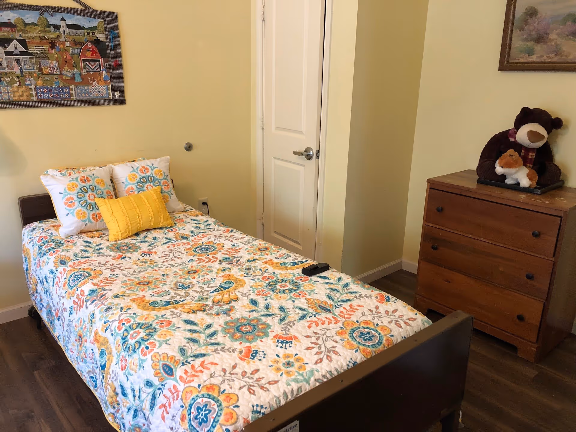A single bed with colorful floral bedding and decorative pillows in a small bedroom with a wooden dresser and stuffed animals.
