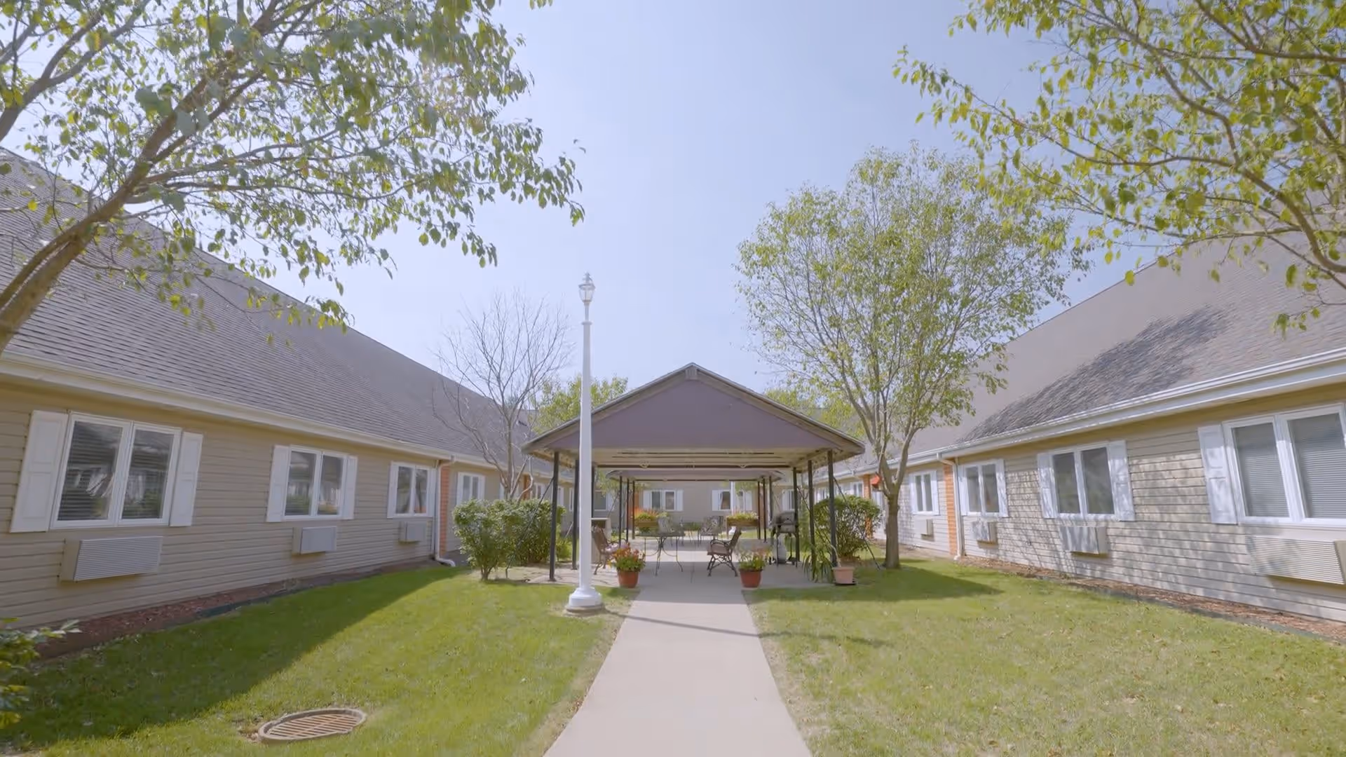 Outdoor courtyard area at Regency Assisted Living with a covered seating area featuring tables and chairs, surrounded by trees and grass, and flanked by beige buildings with white window shutters.