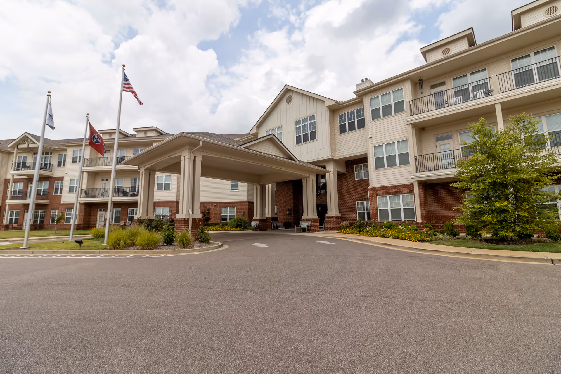 Front entrance of a multi-story senior living building with a covered drop-off porte-cochere, balconies, and flagpoles.